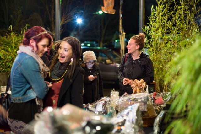 Women engaging and laughing at a table with event items during the Love Bomb 2017 fundraiser for My Voice Music, illuminated by evening lights and surrounded by greenery.